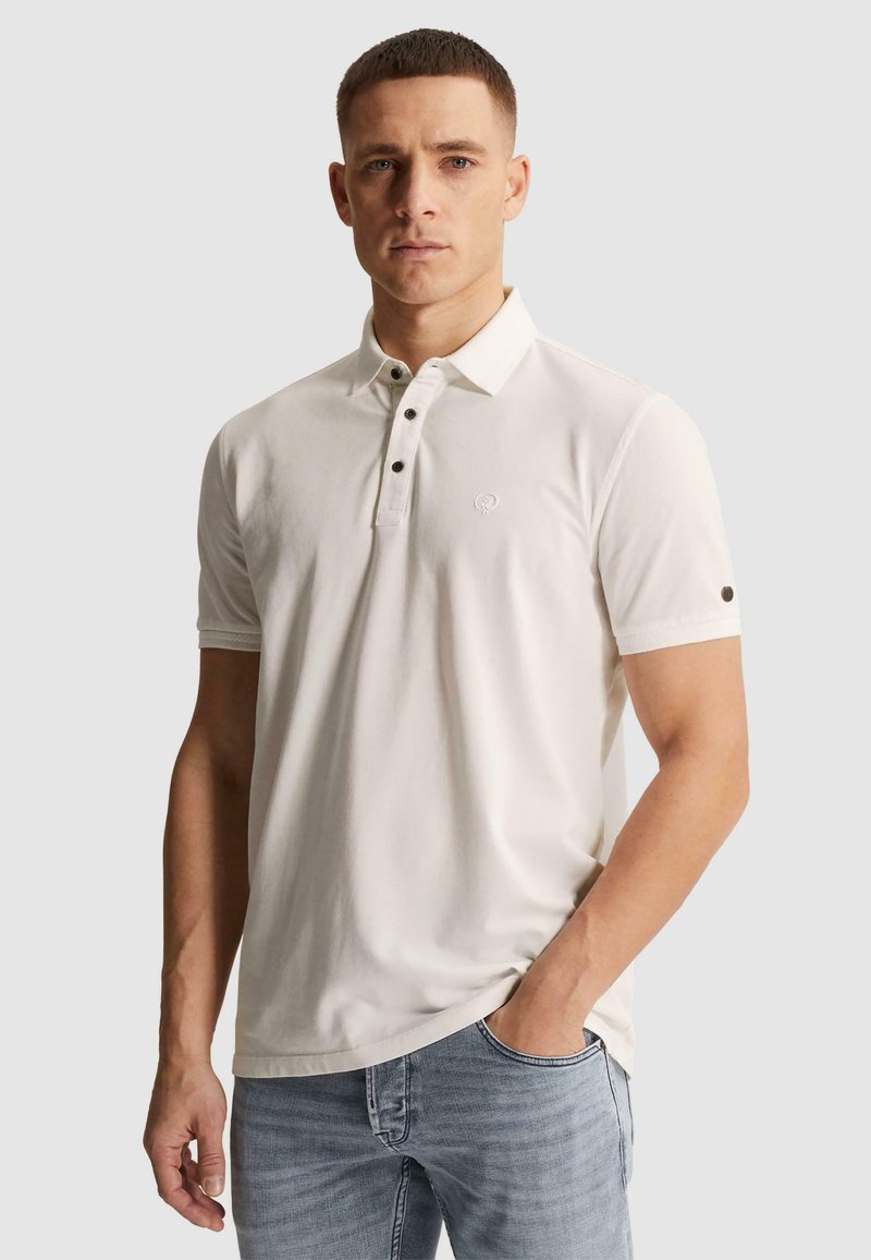 Homme portant un polo blanc à manches courtes avec patte boutonnée et jean bleu clair, main dans la poche, fond neutre.