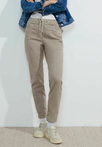 Pantalones beige ajustados con cintura elástica y cordón, bolsillos frontales y textura sutil. Combinados con zapatillas de deporte beige claro y una chaqueta de mezclilla.