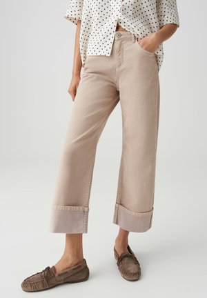 Femme portant un pantalon beige large et court avec de grands revers, un chemisier beige à pois, et des mocassins marron, debout avec une main dans la poche.