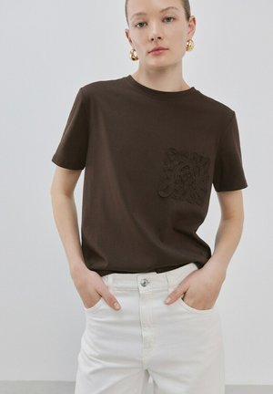 Femme portant des boucles d'oreilles créoles dorées, un t-shirt marron foncé avec un blason brodé sur la poitrine, et un pantalon blanc taille haute, les mains dans les poches.