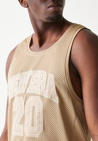 Μπεζ αμάνικη μπλούζα από δίχτυ με λευκούς κεντημένους αριθμούς και το κείμενο "NEW ERA" στο μπροστινό μέρος. Διαθέτει στρογγυλή λαιμόκοψη και φαρδιές μασχάλες.