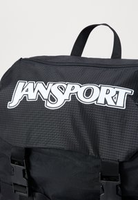 Sac à dos noir avec un motif en grille, présentant un grand logo blanc "JANSPORT", des boucles en plastique et une poignée supérieure pour le transport.