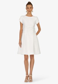Robe blanche à manches courtes avec un corsage ajusté et une accent en dentelle à la taille. Jupe évasée et tissu lisse. Portée avec des sandales blanches à lanières.