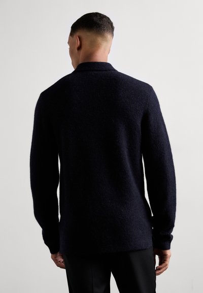 Dunkelblauer Wollpullover mit weicher Textur, langen Ärmeln und Kragen, von hinten vor einem neutralen Hintergrund gezeigt.