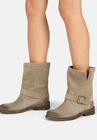 Botas de tobillo de ante beige con una textura trenzada, que presentan un corte en forma de V en el lateral, una correa con un hebilla metálica y un tacón bajo de madera.