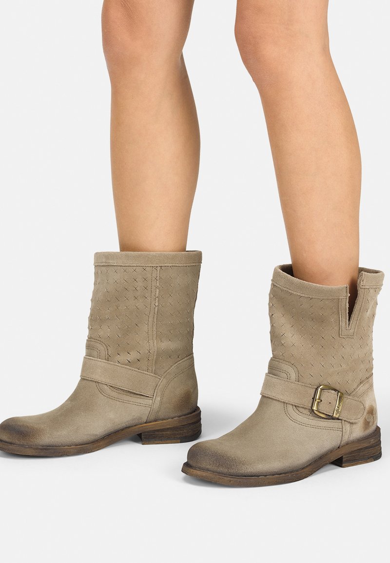 Botas de tobillo de ante beige con una textura trenzada, que presentan un corte en forma de V en el lateral, una correa con un hebilla metálica y un tacón bajo de madera.
