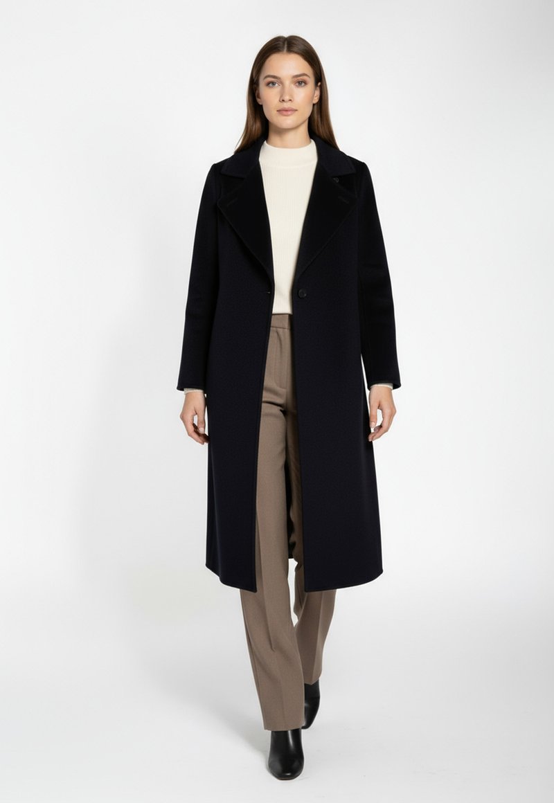 Kvinna som går framåt klädd i en lång svart kappa, krämfärgad polotröja, beige byxor och svarta ankelskor mot en vit bakgrund.