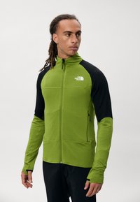 Zielona kurtka zapinana na zamek z czarnymi rękawami, wykonana z teksturowanej tkaniny. Posiada otwory na kciuki, boczne kieszenie oraz logo na klatce piersiowej.