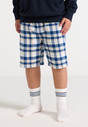 Henkilö pukeutunut sinisiin, beigeihin ja valkoisiin ruudullisiin shortseihin, valkoiset keskipitkät sukat, joissa siniset ja ruskeat raidat, seisoo valkoisella lattialla.