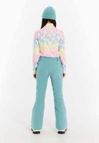 Farebná dlhá košeľa s pravidelným vzorom v štýle tie-dye, doplnená svetlomodrými lyžiarskymi nohavicami. Pletená tyrkysová čiapka. Funkčný dizajn pre zimné aktivity.