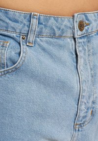 Vaqueros de denim azul claro con una textura suave, que presentan costuras naranjas, un botón metálico de marca y una cremalleras. Diseño sencillo de cinco bolsillos.