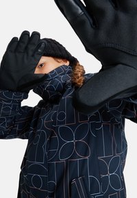 Granatowy płaszcz zimowy w wzór geometryczny, z wysokim kołnierzem i kieszeniami z przodu; w zestawie z czarnymi rękawiczkami i dzianą czapką.