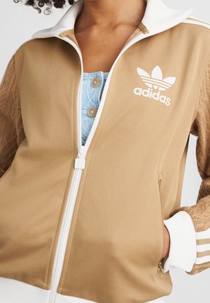Gros plan d'une personne portant une veste zippée beige Adidas avec des bordures blanches et un logo, par-dessus une chemise bleue claire boutonnée.