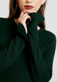 Mörkgrön tröja med långa ärmar, ribbade manschetter och en strukturerad stickad finish. Modellens händer är placerade nära halsringningen.