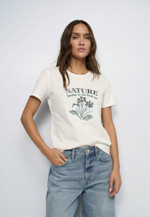 Femme aux longs cheveux bruns portant un t-shirt blanc à motif « "NATURE breathe in the fresh air" » et un jean bleu, debout devant un fond uni.