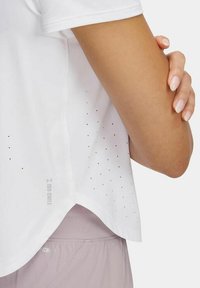 Camiseta deportiva blanca con un dobladillo redondeado, que presenta pequeñas perforaciones y un logo reflectante cerca de la costura lateral. Textura de tela suave.