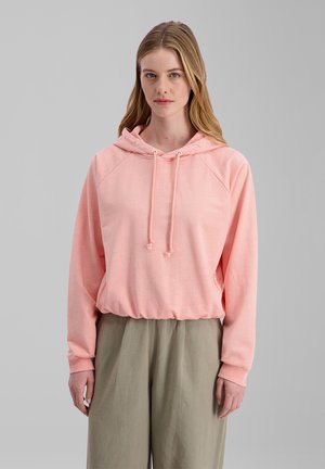 Frau mit langen blonden Haaren, die einen hellrosa Hoodie und lockere beigefarbene Hosen trägt, steht vor einem schlichten grauen Hintergrund.