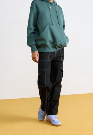 Osoba v zelené mikině Nike, černých kapsáčových kalhotách s bílým prošitím a bílých teniskách s oranžovým logem, stojící na žluté podlaze.