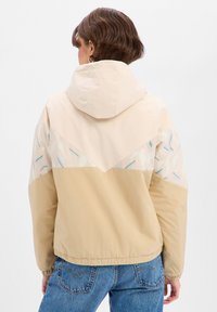 Veste beige légère avec capuche, présentant une partie supérieure à motif géométrique en crème avec des accents bleus, des poignets élastiques et une ceinture ajustée.