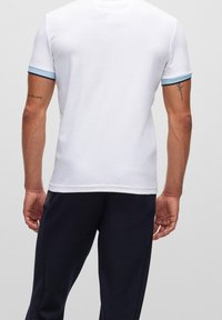 Chemise blanche à manches courtes avec des accents côtelés bleus et noirs au niveau des poignets, présentant une texture lisse et une coupe classique.