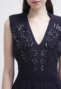 Marinblå klänning med djup V-ringning, prydd med intrikata svarta utsmyckningar och texturerat tyg. Dekorerad med juvel-liknande detaljer och veck.