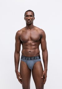 Muž s vypracovaným telom stojí čelom, na sebe má sivé boxerky Calvin Klein s čiernym pásom a logom na jednoduchom bielom pozadí.