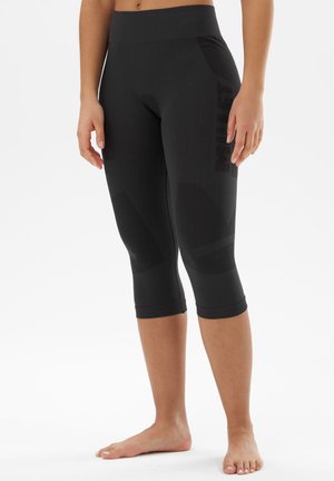 Personne portant un legging de sport noir, ajusté, au genou, debout pieds nus sur un fond blanc.