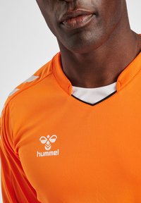 Oranje sportshirt met een v-halsontwerp, witte accenten en een logo op de linkerkant van de borst. De stof lijkt glad en lichtgewicht te zijn.