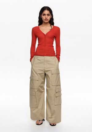 Kobieta stojąca z rękami w kieszeniach, ubrana w długi czerwony top z długim rękawem, beżowe szerokie spodnie typu cargo oraz czarne sandały na obcasie na białym tle.