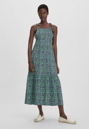 Ärmelloses, blumiges Kleid mit gestuftem Saum, das grüne und lila Muster aufweist. Der Stoff ist leicht, um ihn mit hellfarbigem Schuhwerk zu tragen.