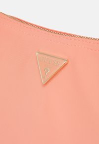 Pochette rose corail avec logo triangulaire doré Guess U.S.A. et une fermeture éclair dorée sur le bord supérieur.