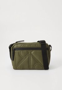 Μικρή ραμμένη τσάντα χιαστί σε ελιά πράσινο χρώμα με μαύρη τσέπη με φερμουάρ και ρυθμιζόμενο μαύρο λουρί σε απλό φόντο.