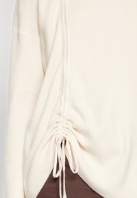 Pull crème côtelé avec détails froncés sur les côtés et cordons, présentant une texture douce et une coupe décontractée, porté sur un haut marron.