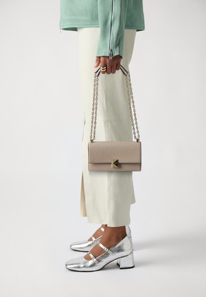 Beige leather handbag with a rectangular shape and a geometric clasp, held by a person wearing silver heels and pale green attire.