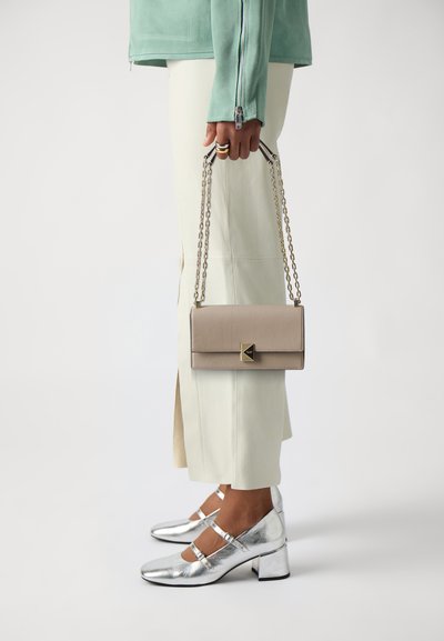 Beige leather handbag with a rectangular shape and a geometric clasp, held by a person wearing silver heels and pale green attire.