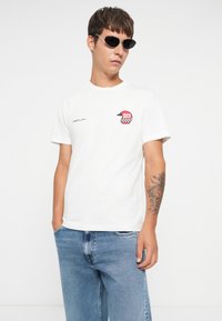 White cotton T-shirt featuring a graphic patch of a helmet and checkered pattern. Paired with light blue denim jeans. Minimalist design.