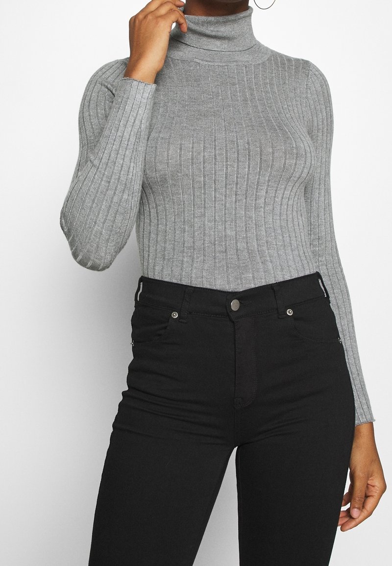 Femme portant un pull col roulé gris côtelé ajusté et un jean noir taille haute, posant avec une main près du visage.