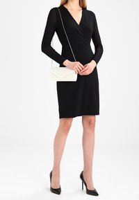 Mujer vestida con un vestido negro ajustado de manga larga, tacones altos negros y sosteniendo un bolso de mano blanco con cadena plateada.