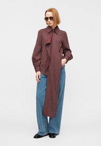 Blouse rayée bordeaux avec un long lien, manches longues et col classique, associée à un jean denim bleu ample et des chaussures noires.