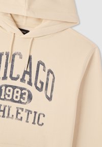 Felpa con cappuccio color beige con una grafica sbiadita in navy che riporta "CHICAGO 1983 ATHLETIC." Presenta cordini e una consistenza di tessuto morbido.