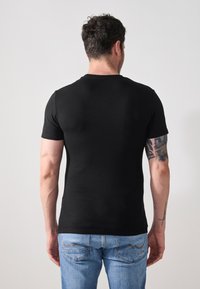 Tricou negru cu mânecă scurtă și guler rotund, textură netedă, formă ajustată, purtat cu blugi albaștri deschiși și tatuaje vizibile pe brațe.