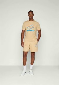 Conjunto de t-shirt e calções em algodão bege com um logótipo colorido da Lotto. O conjunto tem um corte descontraído e é combinado com ténis brancos.
