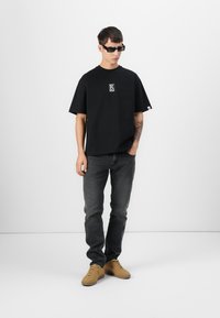 Black oversized t-shirt with a white logo, paired with dark gray jeans and tan suede shoes. Model wearing dark sunglasses.