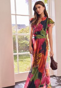 Maxikleid mit floralen Mustern in lebendigen Farben (Pink, Grün, Orange) und Rüschenärmeln, tailliert, kombiniert mit einer braunen Handtasche.