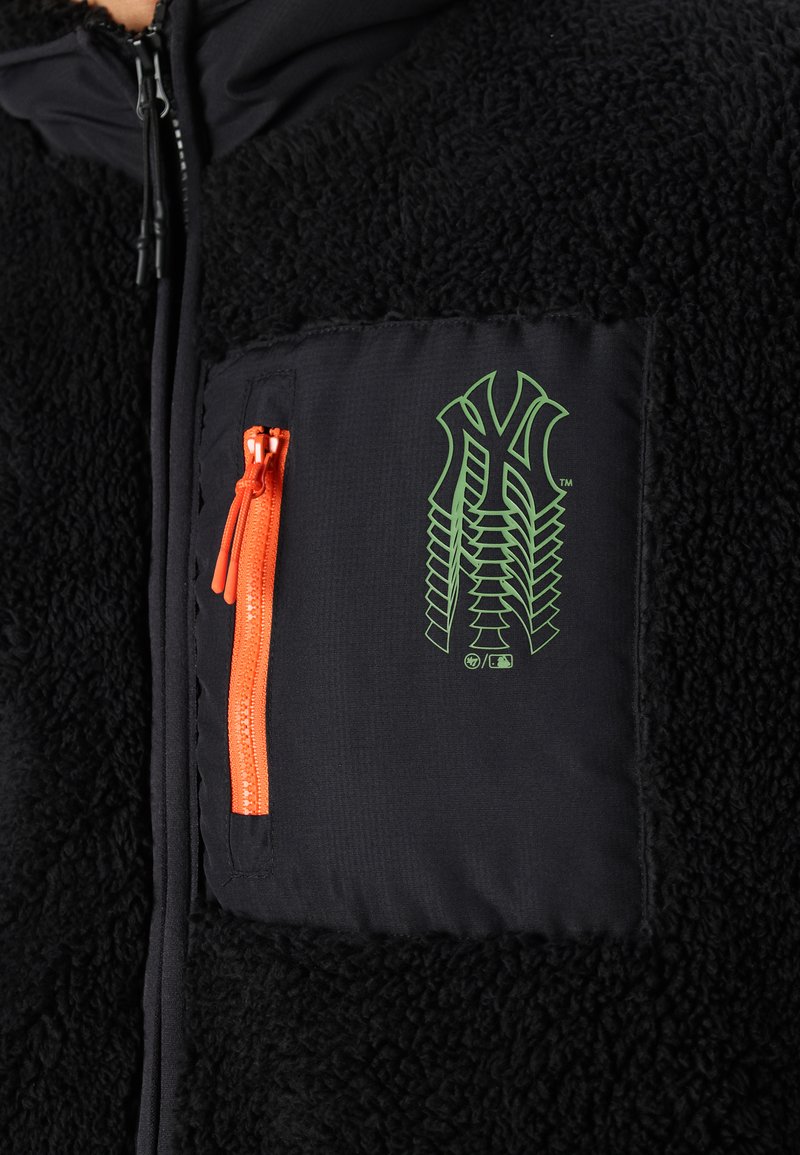 Chaqueta de forro polar negra con un gráfico verde en un bolsillo de tela negra en el pecho. Presenta un zipper naranja en el bolsillo para un cierre seguro.