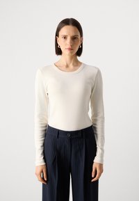 Langermet kremfarget topp i et mykt materiale, med rund hals. Kombinert med marineblå bukser med prikkede striper og dobbel knappelukking.