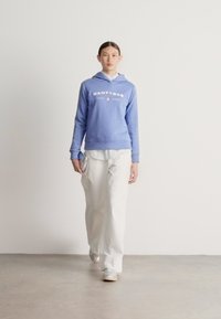 Sudadera con capucha azul claro con texto blanco, sobre una camisa blanca con rayas; combinada con pantalones anchos blancos y zapatillas gris claro. Diseño sencillo.