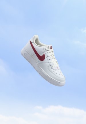 Άσπρο αθλητικό παπούτσι Nike Air Force 1 με κόκκινο λογότυπο Swoosh που αιωρείται πάνω σε φόντο καθαρού γαλάζιου ουρανού.
