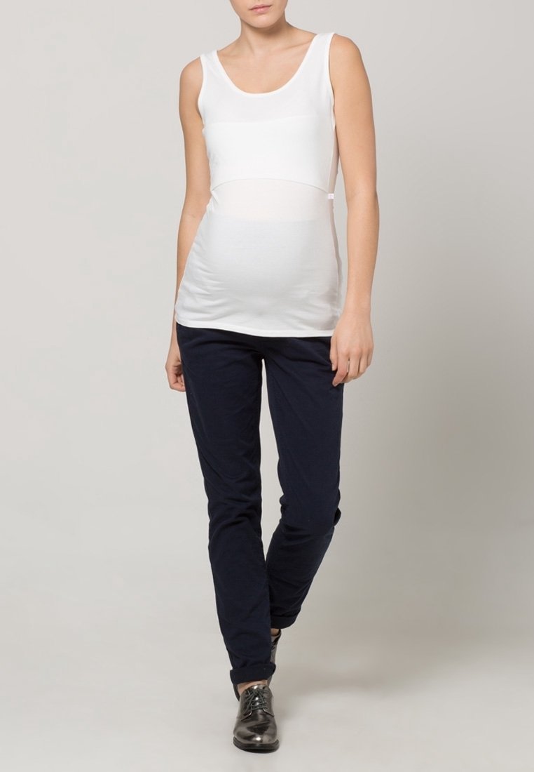 Nainen, jolla on päällä valkoinen hihaton paita ja tummat housut, kävelee eteenpäin vaaleassa yksinkertaisessa taustassa.