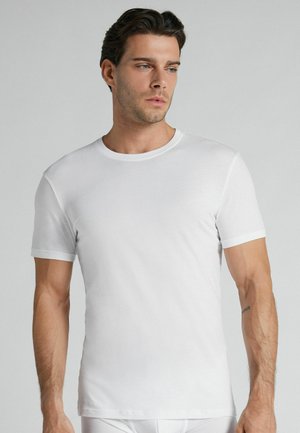 Mand iført en helt hvid t-shirt med korte ærmer og rund hals, står mod en lysegrå baggrund og kigger let til siden.